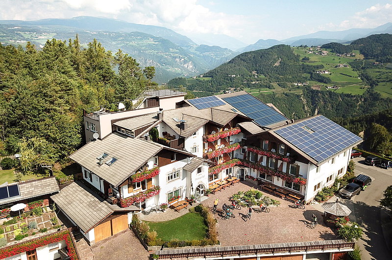 Auf dem Bild ist das Biohotel Steineggerhof von außen gesehen aus der Vogelperspektive. Man sieht die grüne Umgebung aus Südtirol, Italien. Vor dem Bikehotel sind einzelne Fahrradfahrer*innen zu sehen.