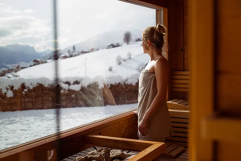 Eine Frau steht in der Saunalandschaft vor einem Panoramafenster mit Blick in die verschneite Bergwelt