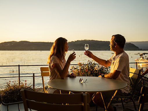 Ein paar sitzt mit Weißweingläsern bei Sonnenuntergang an einem Tisch vor dem Murtensee