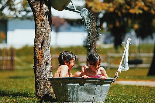 Zwei kleine Kinder sitzen in einer Metallbadewanne auf einer Rasenfläche und werden von Oben mit Wasser besprengt