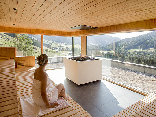 Innenansicht einer großen Sauna mit Panoramafenstern und Blick auf bewaldete Berge und Wiesen
