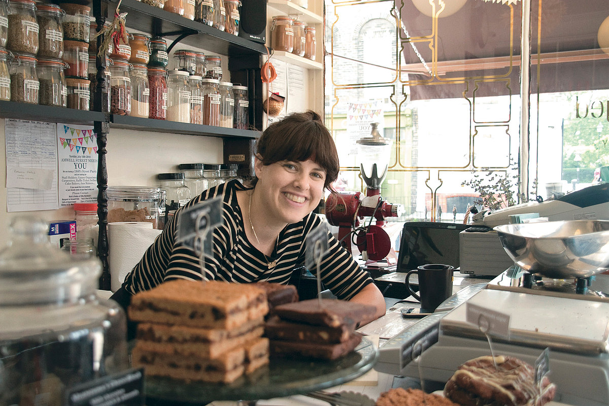 Eine junge Frau steht hinter einen Ladentheke und lächelt in die Kamera