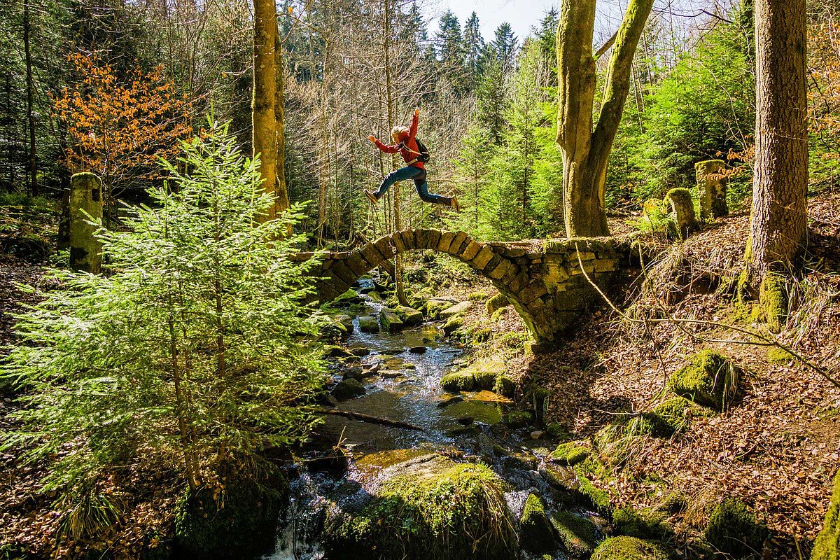 Eine Person spring im Wald über eine kleine, gebogene Steinbrücke