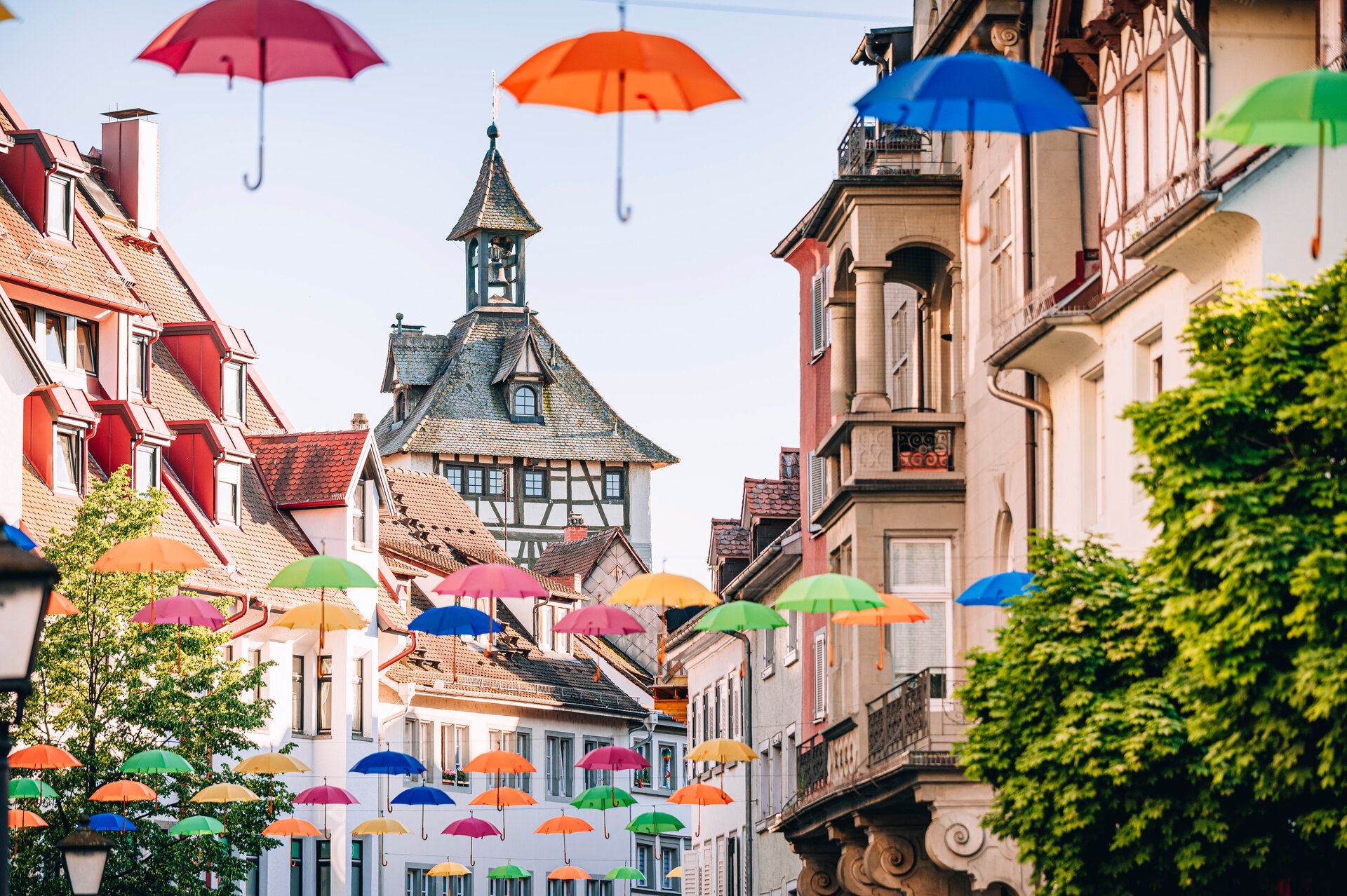 Altstadt-Häuserfassaden, ein Turm im Hintergrund, vom Himmel hängen bunte Regenschirme